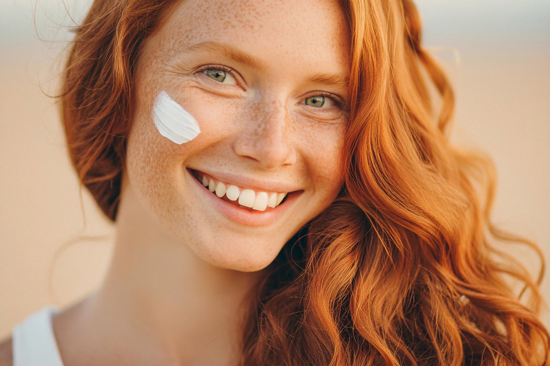 Close-up face of happy long-haired smiling young woman with red hair, freckles and pigmentation on her face with a smear of sunscreen on her skin. Protects the skin from pigmentation and dark spots in the sun.